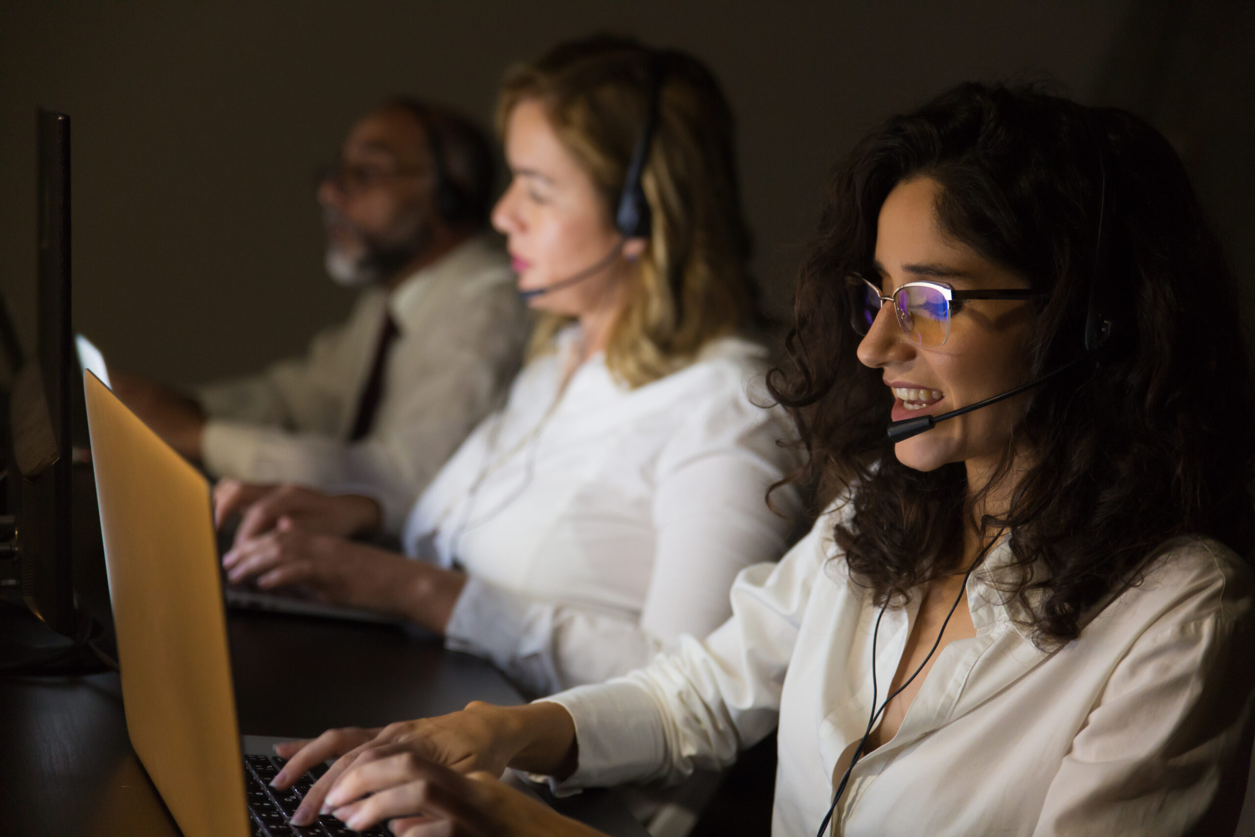 Customer service workers in dark office. Side view of call center operators in headsets using laptops and working late at night. Customer support concept